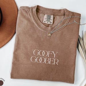 Può includere: T-shirt color espresso con la scritta bianca "GOOFY GOOBER". Una collana d'argento con un ciondolo è appoggiata sulla maglietta. La maglietta ha un'etichetta Comfort Colors.