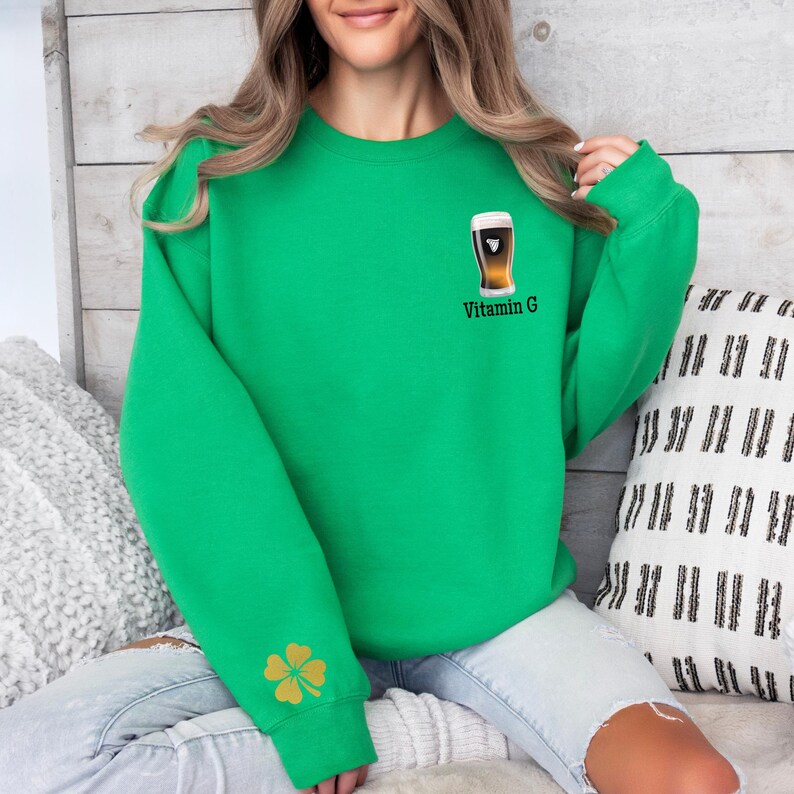 Może przedstawiać: Jasnozielona bluza z małą grafiką szklanki piwa i napisem "Vitamin G" z przodu. Złota koniczyna znajduje się na lewym rękawie. Bluza jest w swobodnym, wygodnym stylu.