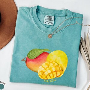 以下が含まれることがあります： 水色のTシャツ。水彩画のマンゴーのデザイン。マンゴーは赤、黄、オレンジ色で、緑の葉が付いています。「MANGO」の文字は黄色で印刷されています。金のネックレスがシャツの上にかけられています。