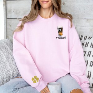 Może przedstawiać: Jasnor&oacute;żowa bluza z nadrukiem kufla piwa i napisem "Vitamin G". Złota czterolistna koniczyna znajduje się na lewym rękawie. Bluza jest w stylu casual.