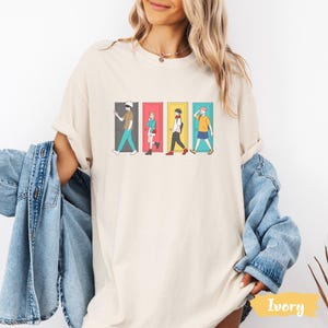 May include: Ivory-colored t-shirt featuring a graphic of four anime characters walking through colorful doors. The doors are gray, pink, yellow, and blue. The characters are in various outfits, with one wearing a blindfold.