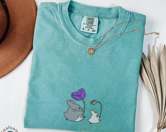 May include: Seafoam green t-shirt with a crew neck and short sleeves. The shirt features a graphic of two cartoon rabbits with a purple flower. A gold necklace with a pendant is draped on the shirt. The tag reads "Comfort Colors".