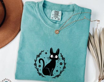 May include: Seafoam green Comfort Colors t-shirt with a black cat graphic inside a floral wreath. The shirt has a crew neck and short sleeves. A gold necklace with a pendant rests on the shirt. The shirt is size S-P-CH.