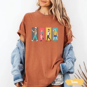 May include: Rust-colored t-shirt featuring a graphic of four anime characters walking through colorful doors. The design includes characters in various outfits, with doors in green, pink, yellow, and blue. The shirt is a casual wear item.