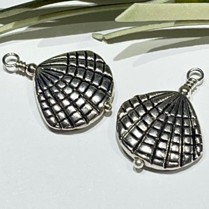 May include: Two silver seashell charms with a textured, grid-like pattern. Each charm has a small loop at the top for attaching to a chain or other jewellery component. The charms are displayed on a white surface.
