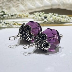 以下が含まれることがあります： 紫色のクリスタルビーズのイヤリング2つ。精巧な銀色の金属細工が施されています。ビーズは光を捉え、金属細工には複雑な円形のデザインがあります。イヤリングには、取り付け用の小さなループが付いています。