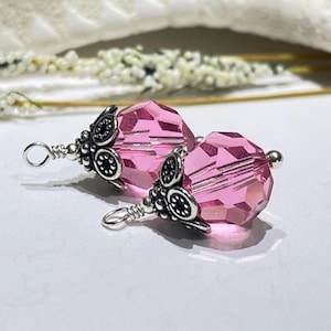 以下が含まれることがあります： 銀色のフィリグリーキャップとループが付いた、2つのピンク色のクリスタルビーズ。ファセットビーズは鮮やかなピンク色で、白い表面に表示されています。フィリグリーキャップは、詳細で装飾的なデザインです。