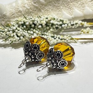 以下が含まれることがあります： 琥珀色のファセットクリスタルビーズが特徴のイヤリングのペア。イヤリングは、フローラルデザインの装飾的なシルバー調の金属細工が施されています。背景には白と緑の花柄のアクセントが含まれています。