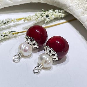 May include: A pair of earrings featuring a deep red, round bead with a silver filigree cap, paired with a white pearl drop. The earrings are set against a white background with floral accents, showcasing a classic jewelry design.