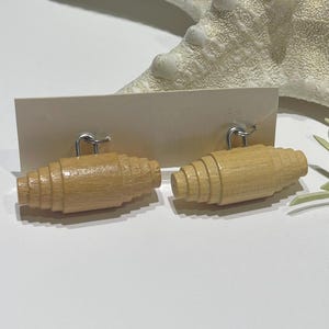 Puede incluir: Un par de pendientes de madera marrón claro con un diseño en forma de barril. Los pendientes están sujetos a una tarjeta de color crema con ganchos plateados. Una estrella de mar blanca y ramitas de plantas verdes están en el fondo.