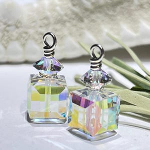 May include: Two clear, iridescent crystal perfume bottle pendants. Each pendant has a square-shaped body and a faceted top with a silver-colored loop. The crystals reflect a rainbow of colors. The background is blurred, with a white surface and green accents.