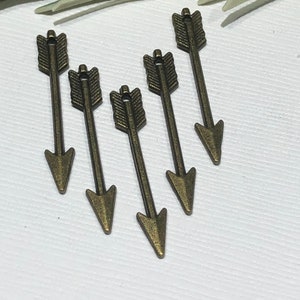 May include: Five bronze arrow charms with detailed feather fletching and pointed tips are arranged on a white surface. The charms are slightly angled, suggesting movement. Greenery is visible in the background.