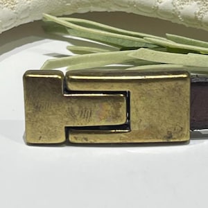 May include: Close-up of a gold-colored metal clasp on a brown leather strap. The clasp is rectangular with a T-shaped design. The background includes a white surface, a textured off-white fabric, and green sprigs.