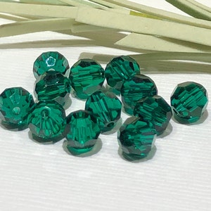 May include: A collection of faceted, emerald green beads. The beads are round with multiple facets that catch the light, creating a sparkling effect. The beads are scattered on a white surface, with a few dried, light green stems in the background.