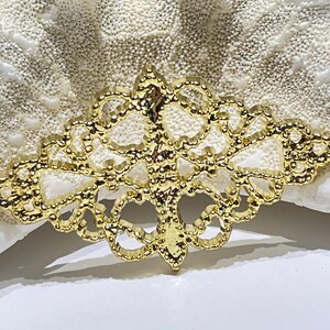 May include: A gold-colored filigree embellishment with an intricate, symmetrical design. The decorative piece has a detailed, openwork pattern and is set against a textured, off-white background. This ornate element is suitable for jewelry or craft projects.