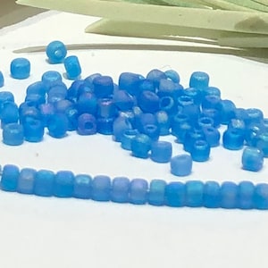 Puede incluir: Un montón de cuentas de semillas iridiscentes de color azul claro, algunas de las cuales están enhebradas en un hilo blanco.