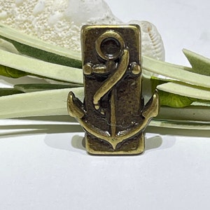 May include: A bronze-colored metal anchor charm with a rectangular shape. The anchor is detailed with a rope loop at the top and a decorative design.
