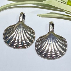 May include: Two silver-toned metal seashell charms with a detailed textured design. The charms have a loop at the top for attaching to jewelry or other crafts.