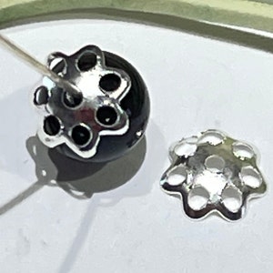 May include: A black bead with a silver-colored floral bead cap. The bead cap has a flower-like design with six circular openings. A needle is inserted through the bead. Another bead cap is next to the bead.