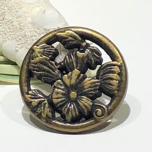 May include: A round, antique gold-toned metal button with a floral design. The button features a raised, detailed design of three flowers with leaves and a swirl.