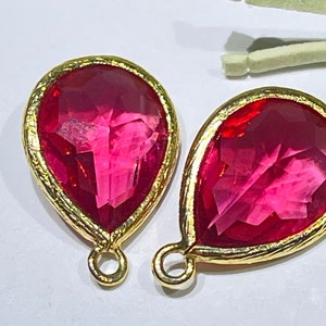 May include: Two pink teardrop-shaped glass stones with gold-toned metal settings. The stones are set in a bezel setting and have a faceted design. The metal settings have a loop at the top for attaching to a jewelry piece.