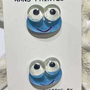 May include: Two hand-painted, whimsical buttons with a blue, cartoon-like design. Each button features two clear, round eyes with black pupils and a smiling red mouth. The card reads "HAND PAINTED" and "CREATIVE BUTTONS BY JHB".