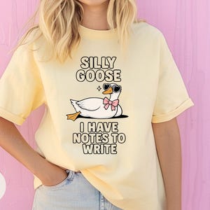 May include: A yellow t-shirt with a cartoon goose wearing sunglasses and a pink bow. The text on the shirt reads "Silly Goose I Have Notes To Write".