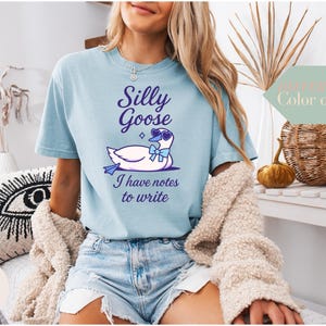 May include: A light blue t-shirt with a white goose wearing sunglasses and a bow. The text on the shirt reads "Silly Goose I have notes to write".