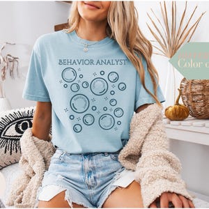 May include: A pale blue t-shirt featuring the words "BEHAVIOR ANALYST" and bubble graphics. The bubbles contain words such as "reinforcement" and "crisis prevention". The shirt is worn with denim shorts and a cream cardigan.