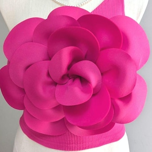 May include: A bright fuchsia one-shoulder top featuring a large, dimensional fabric flower appliqué on the front. The garment is crafted from a ribbed material and is displayed on a mannequin wearing a simple white pearl necklace around the neck.