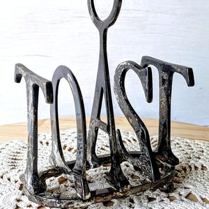 May include: A dark, weathered metal toast rack featuring the word TOAST formed by the upright dividers. The structure has a central carrying loop, a rectangular base with small rounded feet, and a distressed, mottled finish with silver highlights.