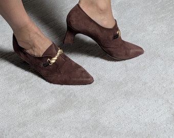 Peut inclure: Chaussures à talons en daim marron avec bouts pointus et détails dorés. Les chaussures ont un petit talon et une chaîne décorative dorée sur le dessus. Les chaussures sont sur un tapis gris clair.