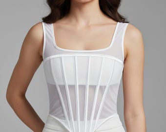 Peut inclure: Un haut style corset blanc doté d'une encolure carrée et de panneaux en maille transparente. Le vêtement est construit avec des canaux de baleines verticaux créant un aspect structuré et lambrissé, se terminant par un ourlet pointu à la taille.