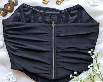 Peut inclure: Haut de corset noir avec un design froncé et une fermeture éclair sur le devant. Le haut est présenté sur une surface blanche avec des boucles d'oreilles dorées et des boutons décoratifs argentés et dorés. Des fleurs blanches sont éparpillées.