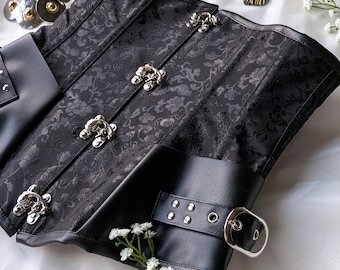 Peut inclure: Corset noir avec motif floral et fermoirs argentés. Le corset présente des détails en cuir noir avec une fermeture à boucle. Des accessoires décoratifs en métal sont visibles en arrière-plan, ainsi que de petites fleurs blanches.