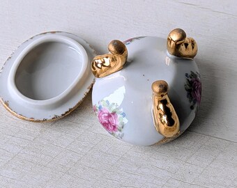 Peut inclure: Petite boîte à bijoux ovale en porcelaine avec couvercle, ornée d'un motif floral de roses roses et de détails dorés. La boîte a trois pieds dorés et une bordure dorée. Le couvercle est ouvert, révélant l'intérieur.