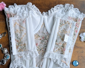 Peut inclure: Corset blanc avec panneaux à motifs floraux et bordures en dentelle. Le corset présente un décolleté en cœur et des baleines pour la structure. Une petite étiquette avec la lettre "S" est visible. Des boucles d'oreilles et des fleurs décorent l'image.