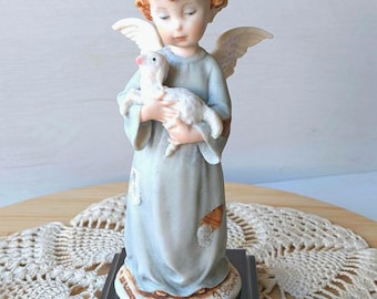Peut inclure: Figurine en céramique d'un ange tenant un agneau. L'ange a la peau claire, les cheveux roux et porte une robe bleu clair. L'agneau est blanc avec un nez rose. La figurine est montée sur une base en bois foncé avec une étiquette dorée "FLORENCE".