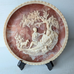 May include: A decorative plate with a relief sculpture depicting a classical scene with figures, a tree, and a lamb. The plate has a reddish-brown background and a cream-colored border. The plate is displayed on a black stand.