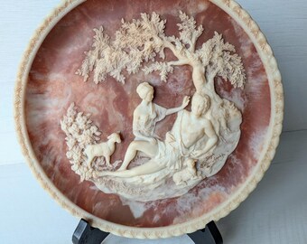 Peut inclure: Une assiette décorative avec une sculpture en relief représentant une scène classique avec des personnages, un arbre et un agneau. L'assiette a un fond brun rougeâtre et une bordure crème. Elle est présentée sur un support noir.