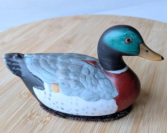 Peut inclure: Figurine de canard en céramique avec un design détaillé. Le canard a une tête turquoise, un cou noir, une poitrine rouge et des ailes grises. Le bec est jaune et la base est noire avec un aspect texturé. La figurine est sur une surface en bois.
