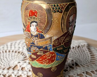 Peut inclure: Un vase en céramique avec une finition craquelée présentant des personnages peints à la main en rouge, bleu et or. Le motif comprend deux personnages distincts avec des auréoles, des motifs complexes et des bordures dorées sur fond brun.