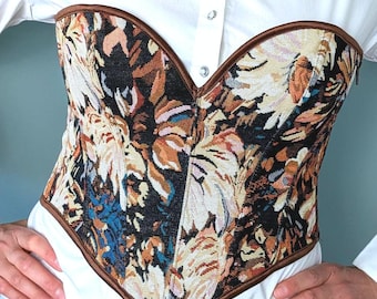 Peut inclure: Un corset en tapisserie florale avec une bordure marron. Le corset présente un mélange de couleurs, dont le noir, l'orange et le bleu. Le vêtement est porté sur une chemise blanche boutonnée.