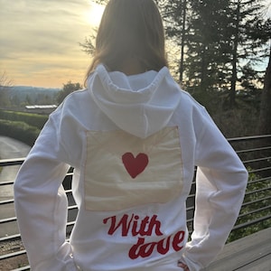 May include: White hoodie with a red heart and envelope design on the back. The words "With love" are embroidered in red script below the graphic. The hoodie has a hood and long sleeves.