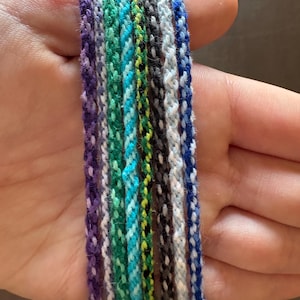 May include: A collection of colorful friendship bracelets in shades of purple, green, blue, and gray. The bracelets are made of woven threads, showcasing a variety of patterns and textures. The bracelets are displayed together, highlighting their diverse color schemes.