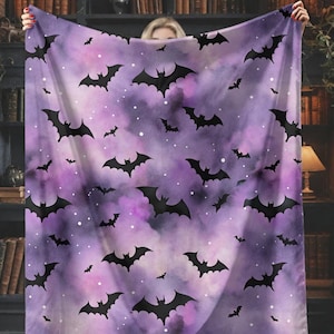 May include: A purple blanket with a pattern of black bats and white specks. The blanket is held up in front of a bookshelf and a black cat is sitting on a windowsill.