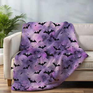 May include: A purple and pink throw blanket with a pattern of black bats. The blanket is draped over a white sofa. The bats are in various sizes and orientations, creating a Halloween theme.