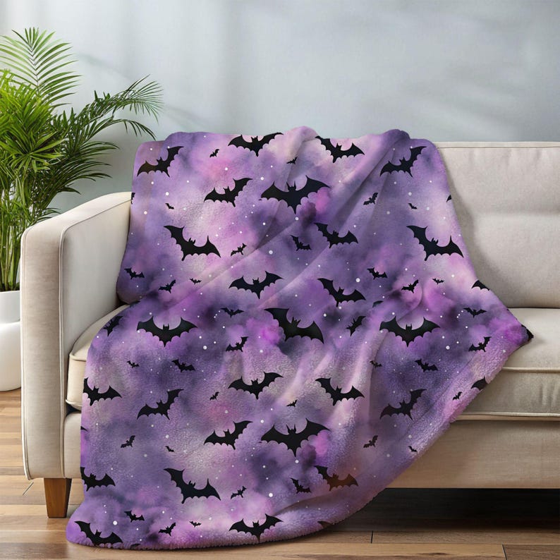 May include: A purple and pink throw blanket with a pattern of black bats. The blanket is draped over a white sofa. The bats are in various sizes and orientations, creating a Halloween theme.