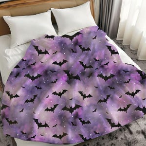 May include: A purple and lavender blanket with a pattern of black bats and white specks. The blanket is draped over a bed with white pillows. The design has a Halloween theme.