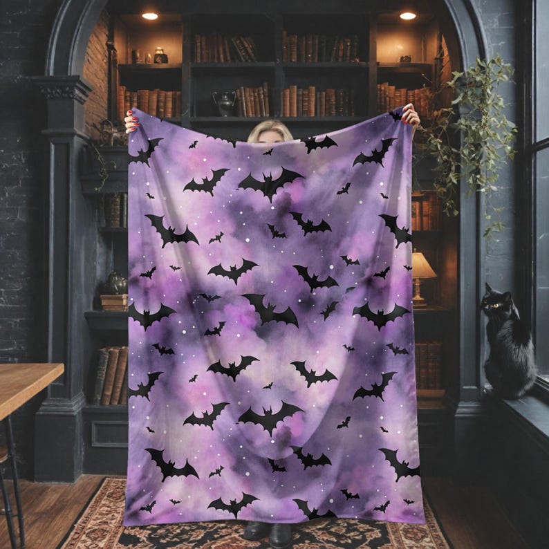 May include: A purple blanket with a pattern of black bats and white specks. The blanket is held up in front of a bookshelf and a black cat is sitting on a windowsill.